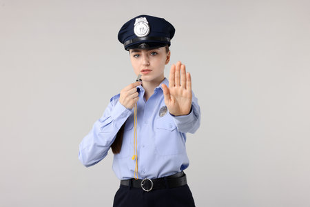 Young policewoman in uniform with whistle showing stop gesture on light gray backgroundの写真素材
