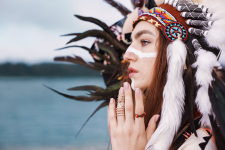 Beautiful shaman woman in native American Indian headdress with face painting and rings outdoors, closeup. Space for textの写真素材