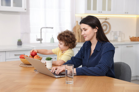 Woman working with laptop while her son playing with apple at wooden desk in home officeの写真素材
