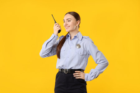 Young policewoman in uniform with walkie talkie on yellow background