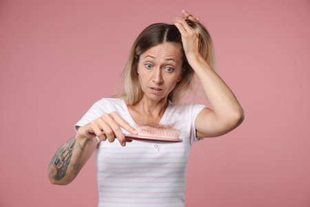 Alopecia problem. Worried woman holding brush with lost hair on pink backgroundの写真素材