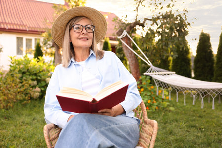 Senior woman reading book in wicker armchair outdoorsの写真素材