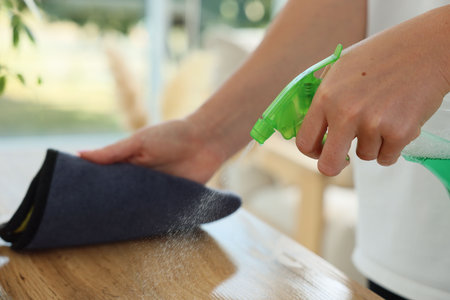 Woman spraying cleaning product while wiping wooden table with rag indoors, closeup