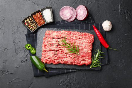 Raw minced meat with rosemary, spices, onion and chili peppers on black textured table, flat lay