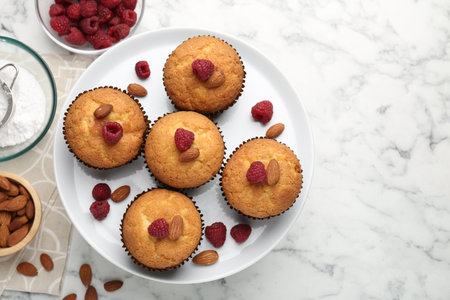 Fresh muffins with raspberries, almonds and flour on white marble table, flat lay. Space for textの写真素材