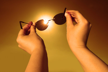 Woman holding sunglasses against orange sky with bright sun, closeup