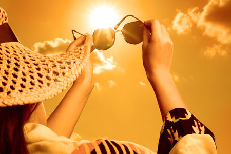 Woman holding sunglasses against orange sky with bright sun, closeupの写真素材