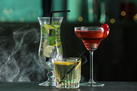 Glasses of tasty cocktails on dark table against blurred background with smoke, bokeh effectの写真素材