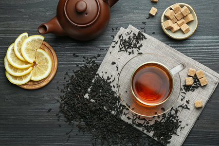 Aromatic black tea in glass cup, dried leaves, brown sugar, lemon and teapot on wooden table, flat layの写真素材