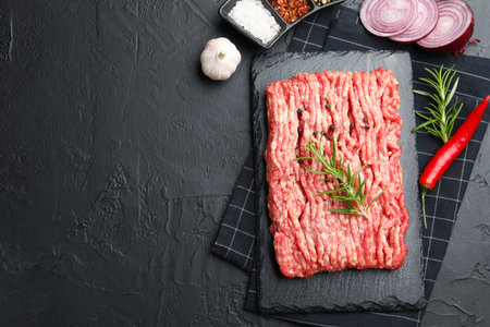 Raw minced meat with rosemary, spices, onion and chili pepper on black textured table, flat lay....