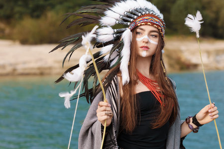 Beautiful woman in native American Indian headdress with face painting, bow and arrow near river outdoors. Space for textの写真素材