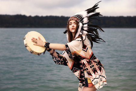 Beautiful shaman woman in native American Indian headdress with face painting and tambourine performing ritual near river outdoorsの写真素材