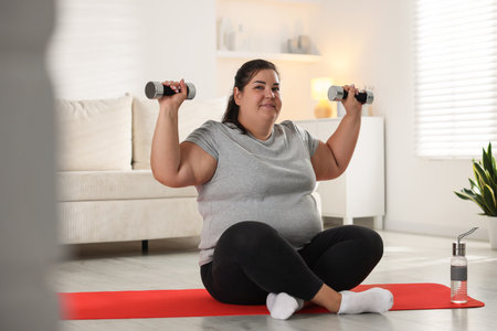 Smiling overweight woman in sportswear training with dumbbells on yoga mat at homeの写真素材