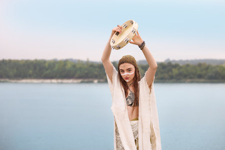 Beautiful woman with face painting and tambourine performing ritual near river outdoors. Space for textの写真素材