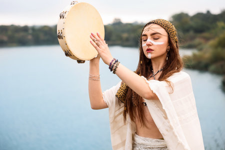 Beautiful shaman woman with face painting and tambourine performing ritual near river outdoors. Space for textの写真素材