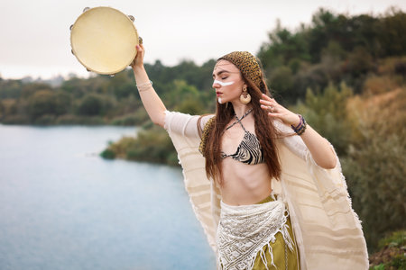 Beautiful shaman woman with face painting and tambourine performing ritual near river outdoors. Space for textの写真素材