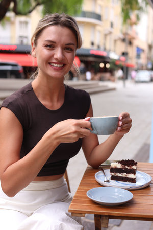 Beautiful young woman with cup of coffee and slice of cake at wooden table outdoorsの写真素材