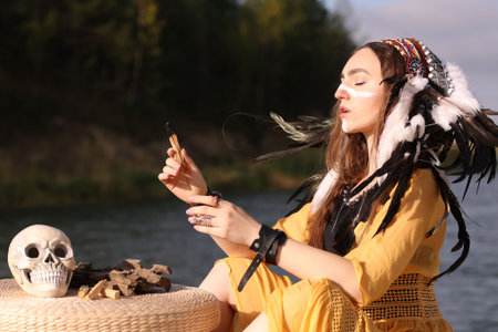 Beautiful shaman woman in native American Indian headdress with face painting, palo santo sticks and skull performing ritual near river outdoorsの写真素材