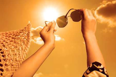 Woman holding sunglasses against orange sky with bright sun, closeupの写真素材