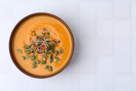 Delicious pumpkin soup with seeds and microgreens in bowl on white tiled table, top view. Space for textの写真素材