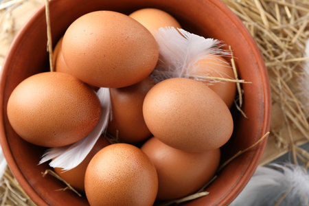 Raw chicken eggs in bowl, feathers and straw on table, top view