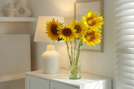 Beautiful sunflowers in vase near lamp on white chest of drawers indoorsの写真素材