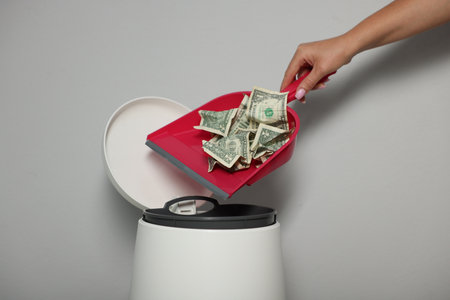 Woman throwing dollar banknotes from dustpan into trash bin near light wall indoors, closeupの写真素材