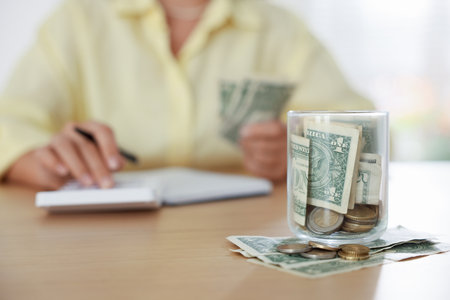 Glass jar with money and woman budgeting at wooden desk, selective focusの写真素材