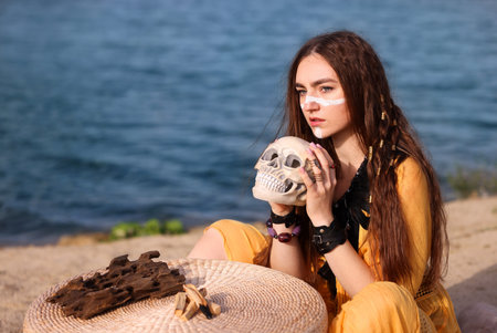 Beautiful shaman woman with face painting, skull and palo santo sticks performing ritual near river outdoors. Space for textの写真素材