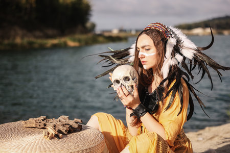 Beautiful shaman woman in native American Indian headdress with face painting, skull and palo santo sticks performing ritual near river outdoorsの写真素材
