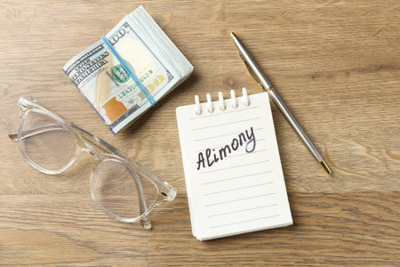 Notebook with word Alimony, money, pen and glasses on wooden table, flat layの写真素材