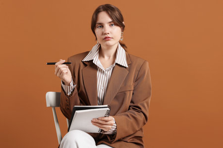 Professional psychotherapist with notebook and pen on chair against brown backgroundの写真素材