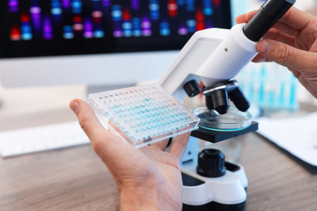 Research in genetics. Scientist working with microscope and microplate at table in laboratory,...