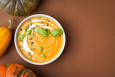 Delicious pumpkin soup with seeds in bowl, basil and vegetables on brown background, flat lay. Space for textの写真素材