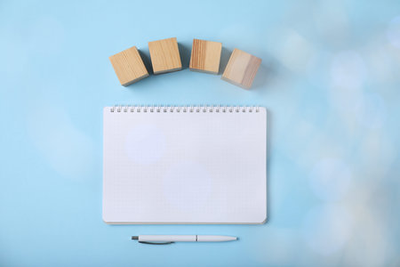 Blank wooden cubes, notebook and pen on light blue background, flat lay. Bokeh effect