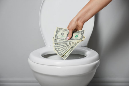 Woman throwing dollar banknotes into toilet bowl near light wall indoors, closeupの写真素材
