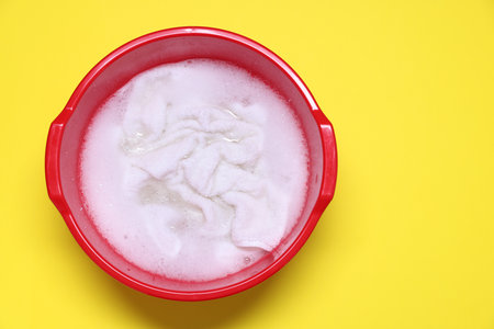 Plastic basin with clothes, water and foam on yellow background, top view. Space for textの写真素材