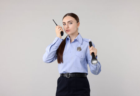 Young policewoman in uniform with flashlight and walkie talkie on light gray backgroundの写真素材