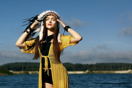 Beautiful shaman woman in native American Indian headdress with face painting near river outdoors. Space for textの写真素材
