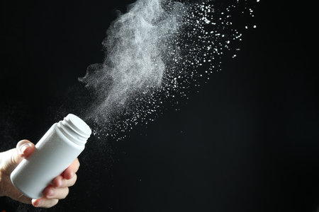Person holding talcum powder bottle with powdery splash on black background, closeup. Space for textの写真素材