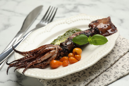Tasty cooked squid served with sauce, tomatoes, cutlery and basil on white marble table, closeup