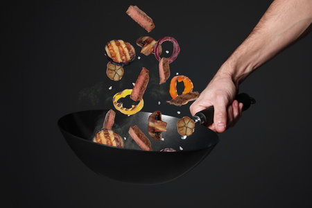 Chef tossing meat and vegetables pieces in cooking pan against black background, closeupの写真素材