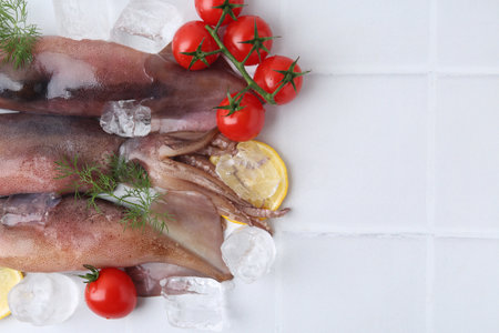 Raw squids, ice cubes, tomatoes, lemon slices and dill on white tiled table, flat lay. Space for textの写真素材