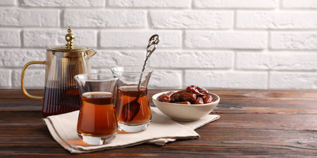 Tasty Turkish tea in glass cups, dates and teapot on wooden table against white brick wall, closeup. Space for textの写真素材