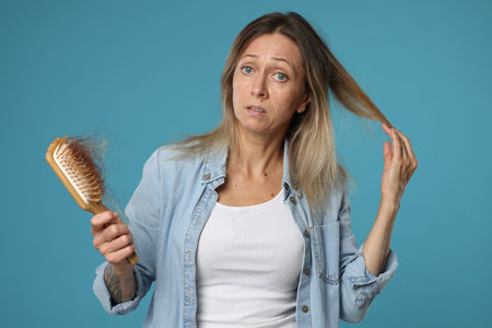 Alopecia problem. Worried woman holding brush with lost hair on light blue backgroundの写真素材