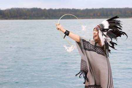 Emotional woman in native American Indian headdress with face painting, bow and arrow near river...