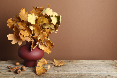 Autumn composition with vase of dry leaves and acorns on wooden table against brown background. Space for textの写真素材