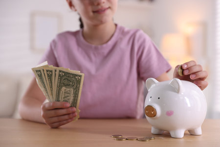 Little girl putting coin into piggy bank at wooden table indoors, closeup. Pocket money and responsibilityの写真素材