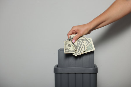 Woman throwing dollar banknotes into trash bin near light wall indoors, closeup. Space for textの写真素材