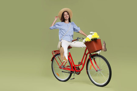 Smiling woman in hat riding bicycle with basket of flowers on green background. Space for text